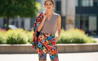 Mladá žena v kvetinových nohaviciach a neutrálnej blúzke drží bundu na pleci, vonku v mäkkom svetle.